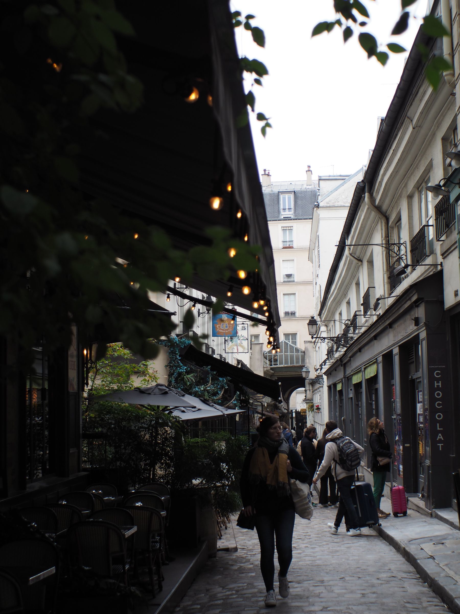 A narrow cobblestone street lined with cafés, people strolling past.