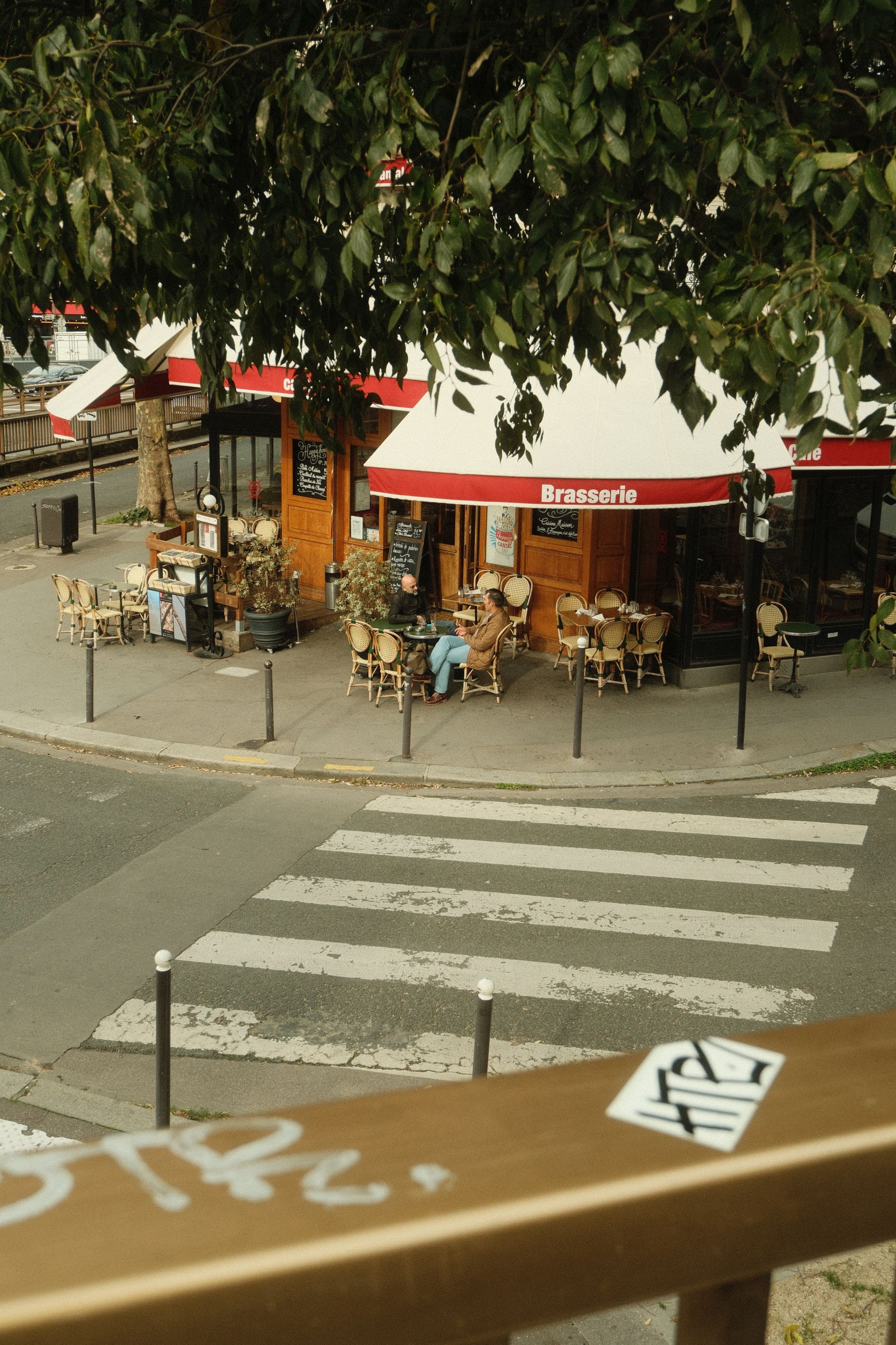 A quiet Parisian street corner, two friends in conversation at a corner brasserie beneath the trees.